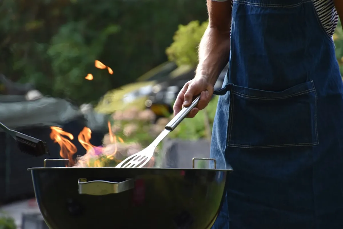 Person i förkläde grillar över öppen låga på en klotgrill i en trädgård i kvällssol